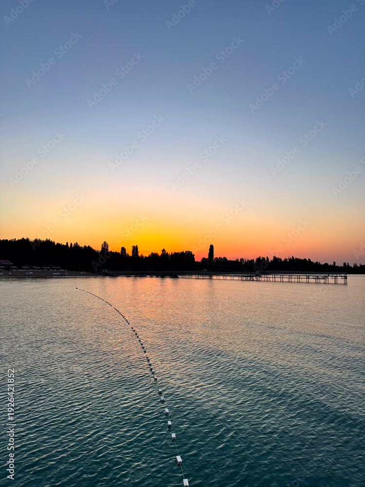 Fototapeta premium Early sunrise at Issyk Kul Lake in Kyrgyzstan. Warm golden sky reflecting in calm water with silhouettes of trees and pier on horizon.