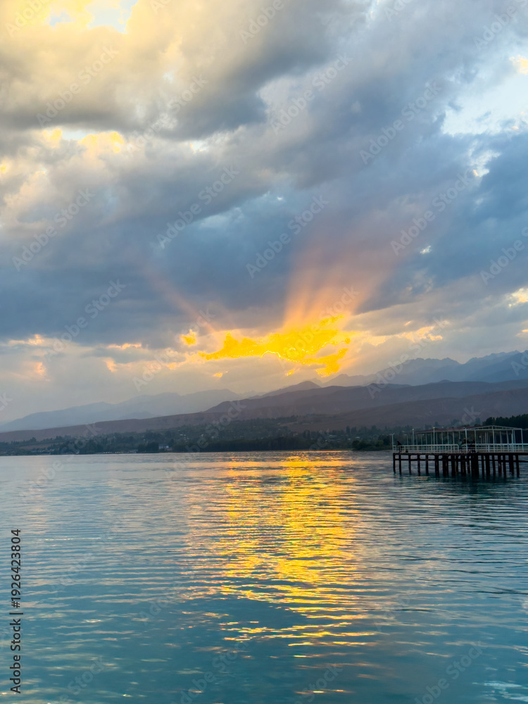Fototapeta premium Sunset at Issyk Kul Lake in Kyrgyzstan. Bright sun breaking through clouds reflecting in clear turquoise water.