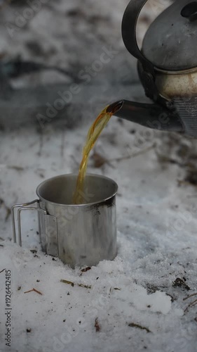 Hot tea is poured from a camping kettle into a metal mug. A hot drink on a winter hike. The concept of bushcraft and wilderness survival.