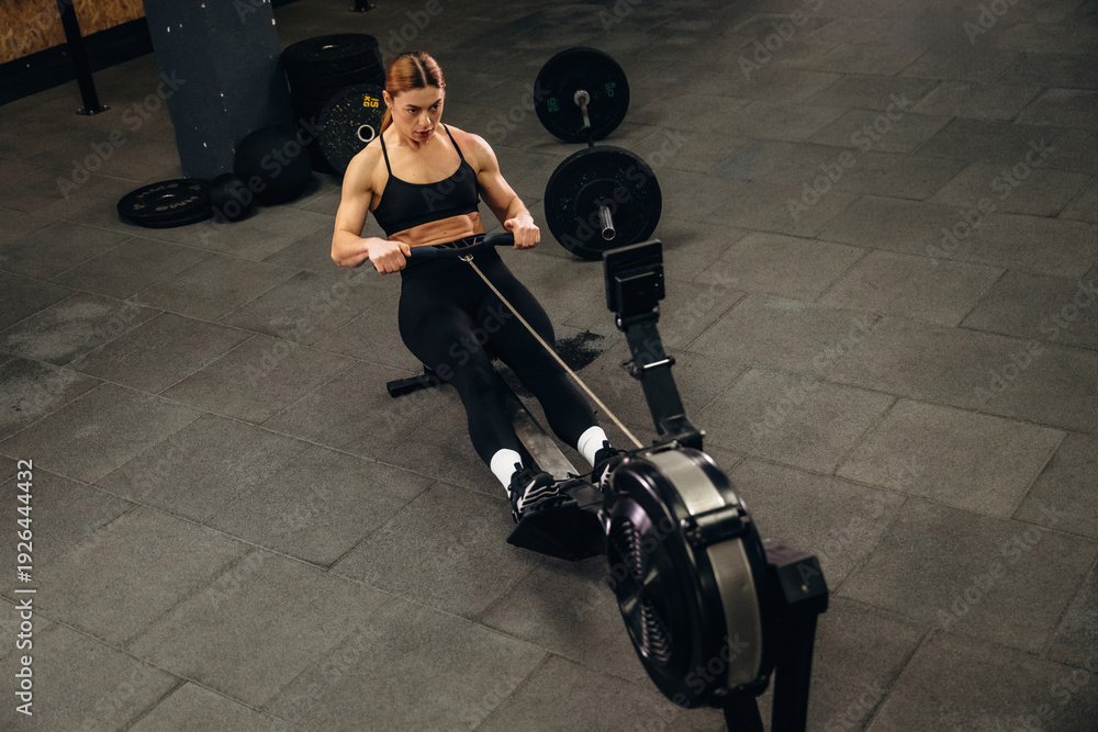 Obraz premium Female athlete performing rowing exercise on machine in gym, with weights and fitness equipment visible in the background