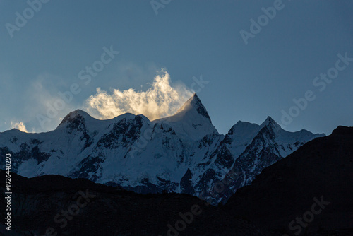 View of jagged, snow-capped peaks pierce the sky, their summits veiled in ethereal clouds touched by the sun's glow, Hunza Nagar, Gilgit, Pakistan.