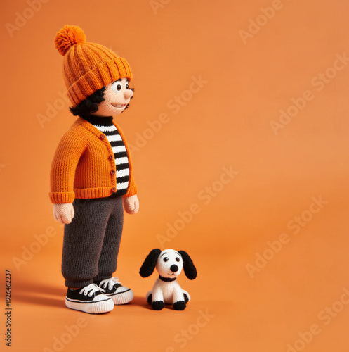Knitted boy doll wearing orange hat stands beside small puppy
