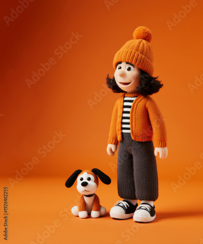 Knitted boy doll wearing orange hat stands beside small puppy