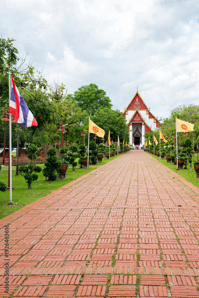 Obraz premium View of the paved ceremonial path leading to the Wihan Phra Mongkhon Bophit in Ayutthaya, Thailand. The path is lined with green bushes and flagpoles flying the Thai national flag and yellow flags.