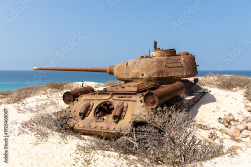View of an old, rusted tank silently rests on the sandy dunes against the backdrop of the turquoise sea and clear blue sky, Qalansiyah, Socotra, Yemen.