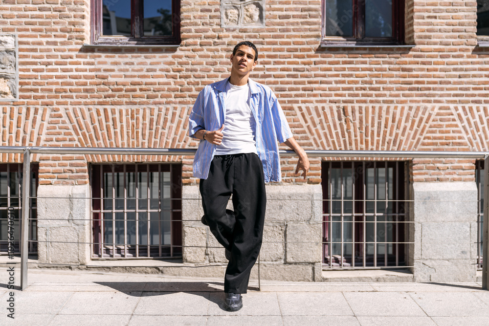 Fototapeta premium Young man in casual streetwear leaning on a railing by a brick building, exuding modern urban style, confidence and youth culture
