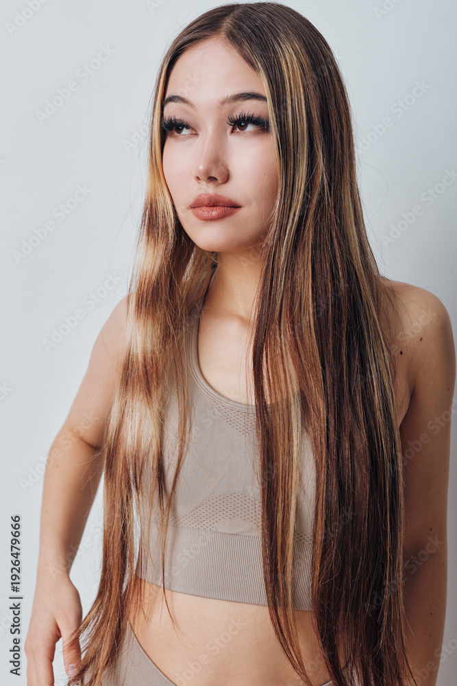 Naklejka premium Young woman with long straight hair and neutral makeup wearing beige crop top standing against light background with serious expression looking aside.