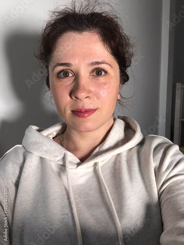 Selfie portrait of woman in cozy hoodie relaxing at home. Natural portrait in a modern domestic setting. Comfort in simplicity, everyday authenticity and serene domestic life concept