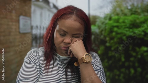 Woman rests cheek on hand wearing wooden watch in front of brick building beside hedge under daylight; weariness.