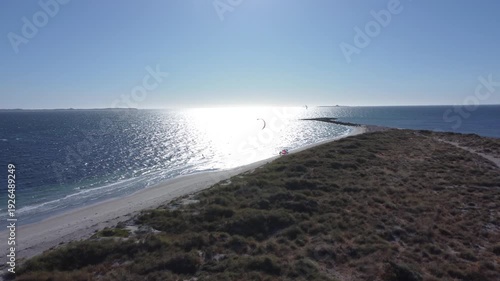 Stunning aerial perspective of a coastal landscape with kitesurfers gliding across the sparkling blue ocean on a bright sunny day, showcasing a popular water sport and beautiful scenery