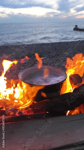Pot of water boiling vigorously on a vibrant bonfire while camping, preparing a hot meal outdoors on a hiking adventure, surrounded by logs and bright yellow flames