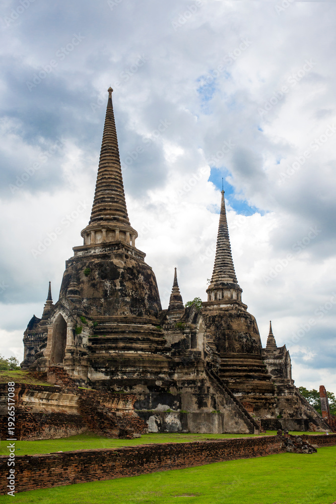 Obraz premium The three iconic restored chedis (stupas) at Wat Phra Si Sanphet in Ayutthaya, Thailand. The massive, bell-shaped structures stand in a row on a raised platform covered in green grass. Cloudy day.