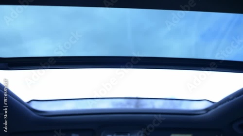A cozy car interior reveals a vast stretch of blue sky and fluffy clouds above, inviting a sense of freedom and peace during a relaxing drive. Opening the car's panoramic sunroof.