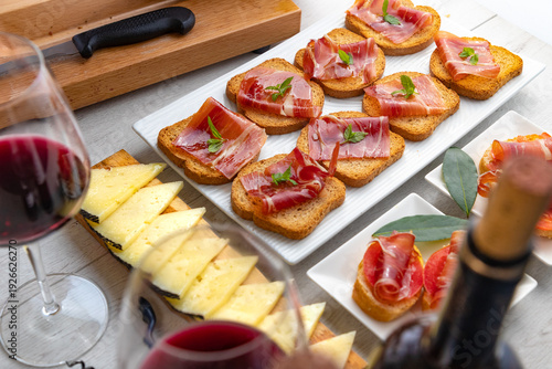 Tapas españolas con jamón, queso y vino tinto.Variedad de tapas españolas con pan tostado, jamón curado, lonchas de queso y copas de vino tinto sobre mesa rústica. Presentación elegante y concepto 