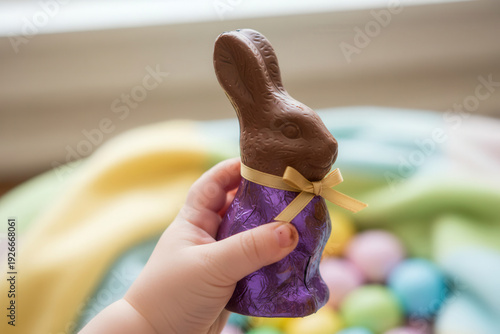 Toddler’s hand holding a chocolate Easter bunny wrapped in purple foil, festive spring holiday concept with colorful eggs in soft focus background, childhood joy, celebration, and seasonal sweets