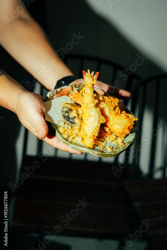 Close-up of a person holding a rustic ceramic plate filled with crispy Japanese tempura, including golden shrimp and assorted vegetables. Dramatic warm lighting enhances the crunchy texture and vibran