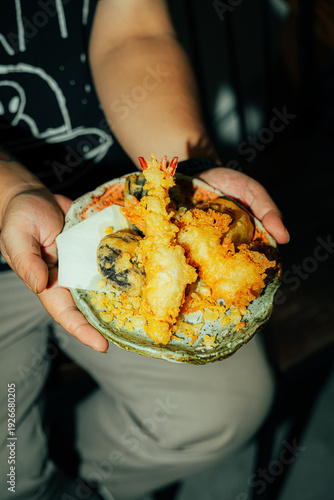 Close-up of a person holding a rustic ceramic plate filled with crispy Japanese tempura, including golden shrimp and assorted vegetables. Dramatic warm lighting enhances the crunchy texture and vibran