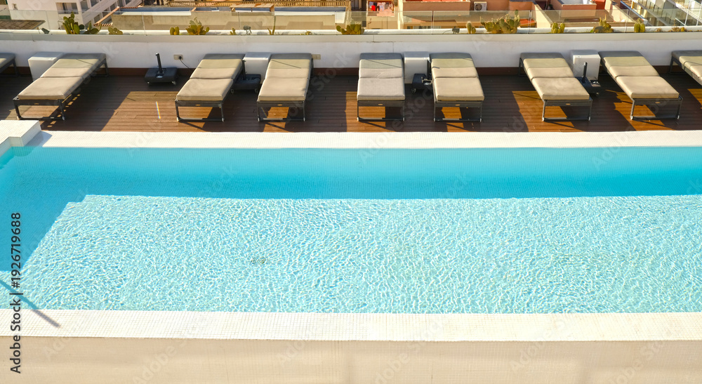 Obraz premium Sunlit rooftop hotel pool with sparkling turquoise water and empty loungers. For travel, real estate, wellness, hospitality and lifestyle branding.