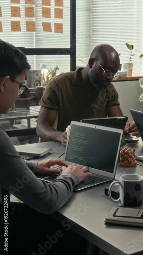 Vertical shot of two diverse web engineers programming at wooden desks during workday in modern office of tech company