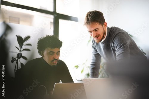 Two men collaborate on a project, looking at a laptop screen in a modern office setting.