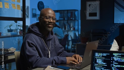 Portrait of Black millennial QA engineer wearing hoodie smiling at camera while testing software product in dimly lit office