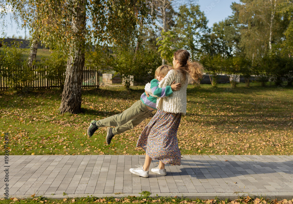 Fototapeta premium An older sister spins her younger brother around, holding his armpits and spinning around in the park. Children are having fun outdoors, enjoying the moment of joy. Life energy. Dynamic photo