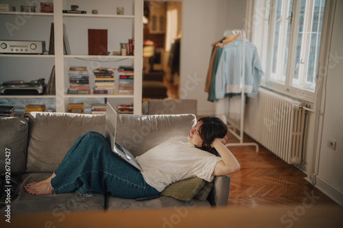 Happy brunette woman stretching and smiling while lying on sofa with laptop in sunny living room
