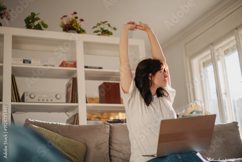 Happy brunette woman stretching with pleasure while working on laptop on sofa in sunny living room
