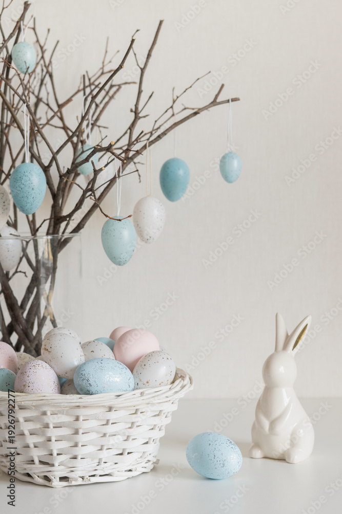 Obraz premium Easter tree with pastel speckled eggs hanging on spring branches in glass vase. White ceramic bunny and basket with painted eggs on light background. Soft natural light, minimal home decor