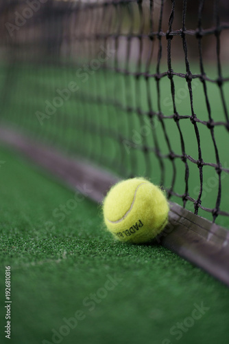 Padel ball resting against a black net on a green court. Close-up of racket sports equipment during a match.