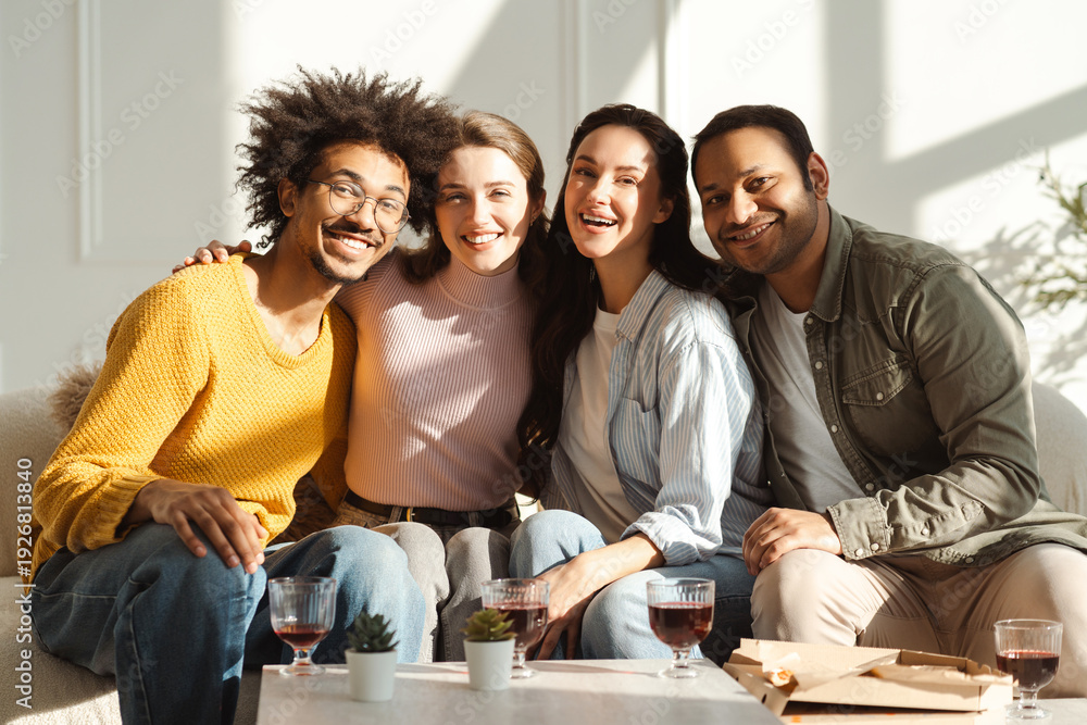 Fototapeta premium Happy smiling diverse multinational friends hugging looking at camera sitting on sofa in living room