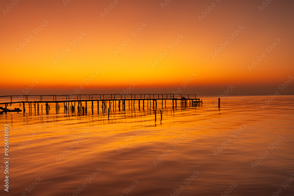 Obraz premium Golden Hour Silhouette of a Wooden Pier over Calm Ocean Water,A serene and minimalist landscape featuring a long wooden pier silhouetted against a vibrant orange sunset. The calm sea perfectly.