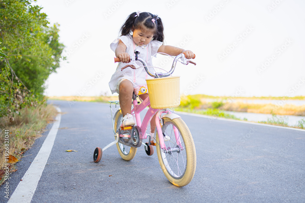 Fototapeta premium Little girl learning to ride a pink bicycle with training wheels on a park road,A cute young Asian girl in a white shirt enjoys riding her pink bicycle with training wheels on an asphalt road surround