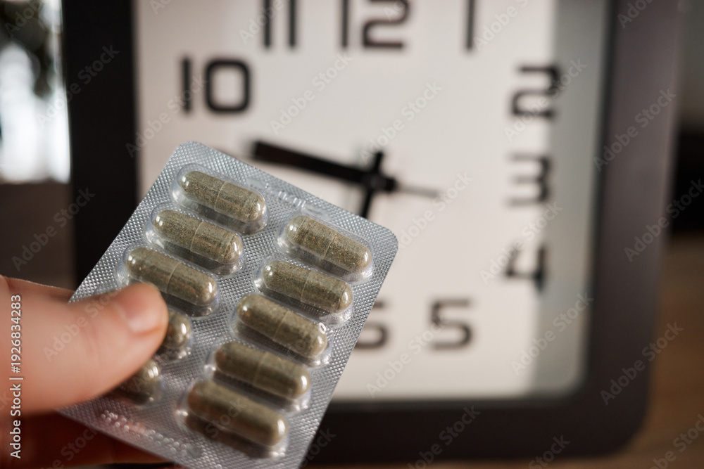 Obraz premium Man holds medicine in hand next to clock showing time for taking pills in a home setting during daylight