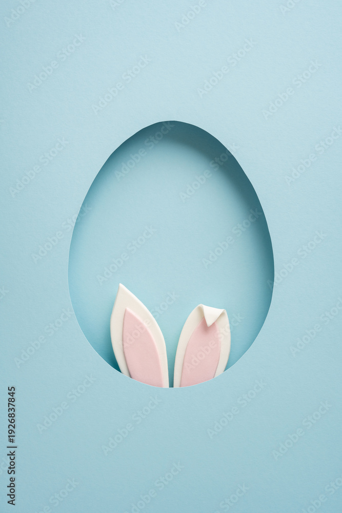 Fototapeta premium Minimal vertical pastel Easter greeting featuring a paper egg cutout with pink bunny ears peeking through, conveying playful spring celebration and charming holiday decoration
