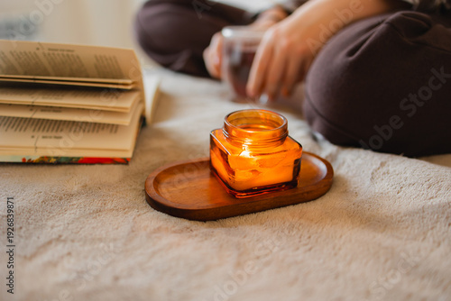 Open book and burning candle on wooden tray over soft blanket. Intimate home setting with warm light. Concept of evening routine, relaxation, slow living and cozy autumn mood.