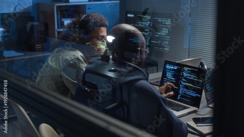 View through glass wall of two multiethnic young software developers discussing bugs both looking at laptop screen displaying code in modern office