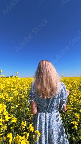 Cinematic FPV drone shot of woman walking through blooming yellow rapeseed field on sunny spring day. Peaceful countryside lifestyle scene for nature, freedom, and slow living themes.