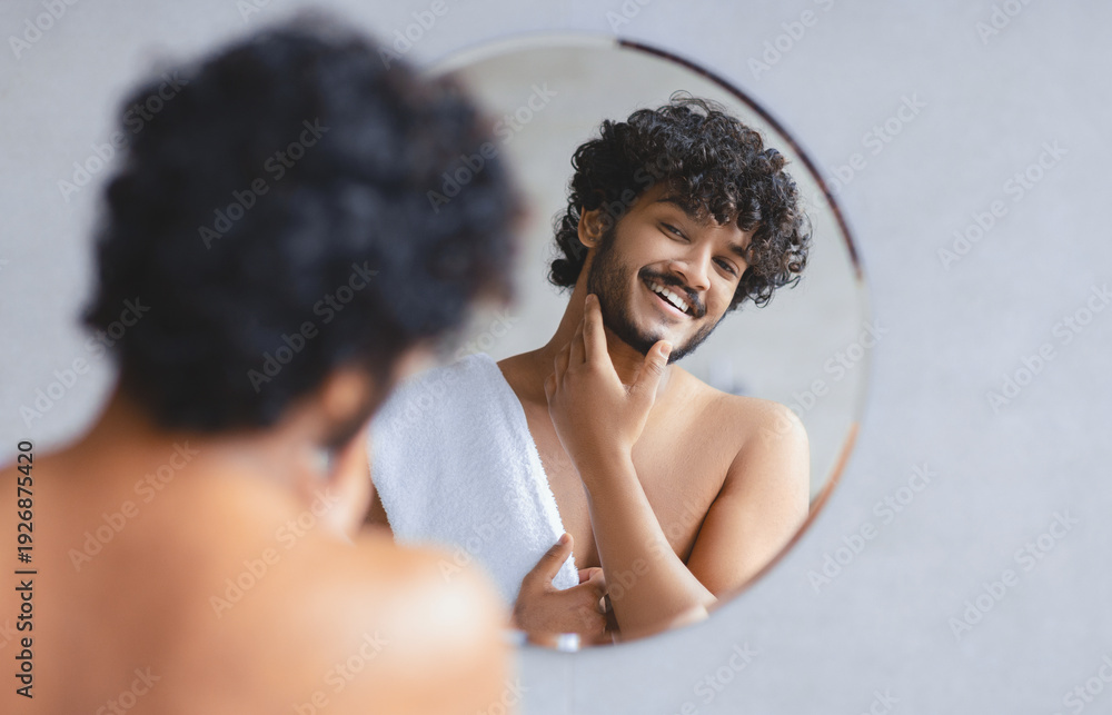 © Prostock-studio - A young man stands in front of a mirror in a bathroom. He is smiling while grooming himself. He has curly hair and is wearing a towel. Morning light fills the room.