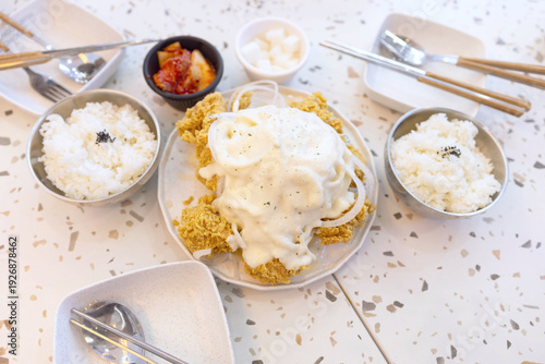 A korean snow onion fried chicken . A plate of creamy white sauce and sliced onions on crispy fried golden breaded chicken sits at the center, surrounded by bowls of steamed rice and kimchi.