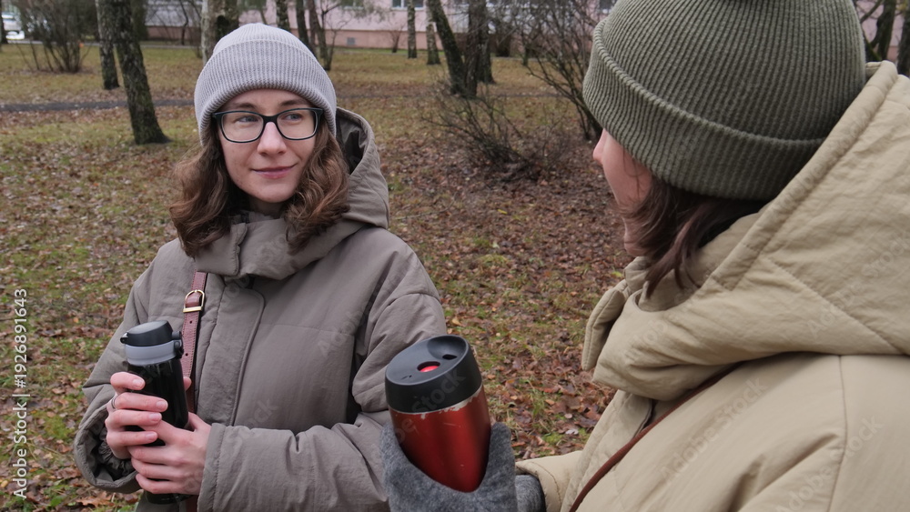 Fototapeta premium Two young women friends spending time together outdoors, sharing a warm drink from thermal mugs while having a private conversation in an autumn park setting