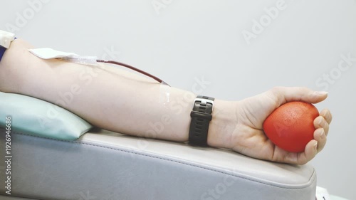 Close-up of a person squeezing a red stress ball while donating blood, with medical tubing and collection equipment visible in a clinical healthcare setting.