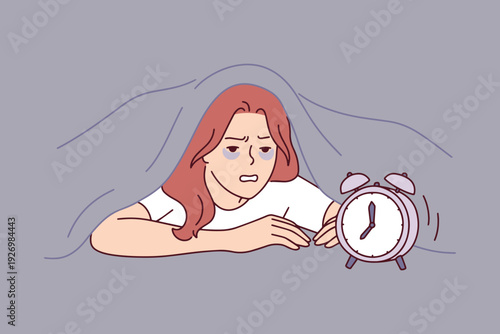 Tired woman wakes up irritated at 7 am from loud alarm clock under blanket. Young girl feels exhausted, annoyed, and lacks sleep after stressful night, struggling to get out of bed.