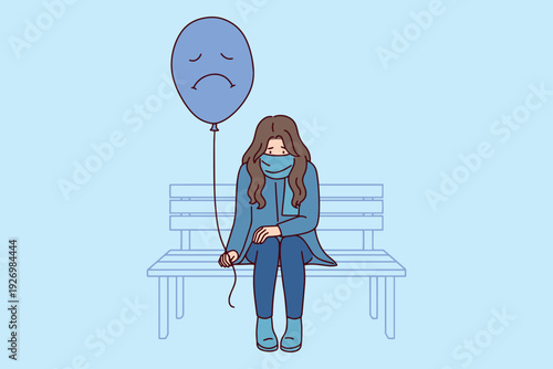 Sad woman sits on outdoor bench holding blue balloon with frowny face during winter or autumn. Concept of Blue Monday, loneliness, depression, and Mental Health struggles in cold season.