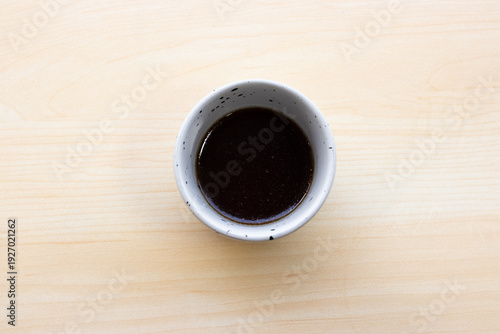 Top view of Japanese noodle dipping sauce or Tsuyu in a small ceramic cup, served on a light wooden background for Asian cuisine themes.