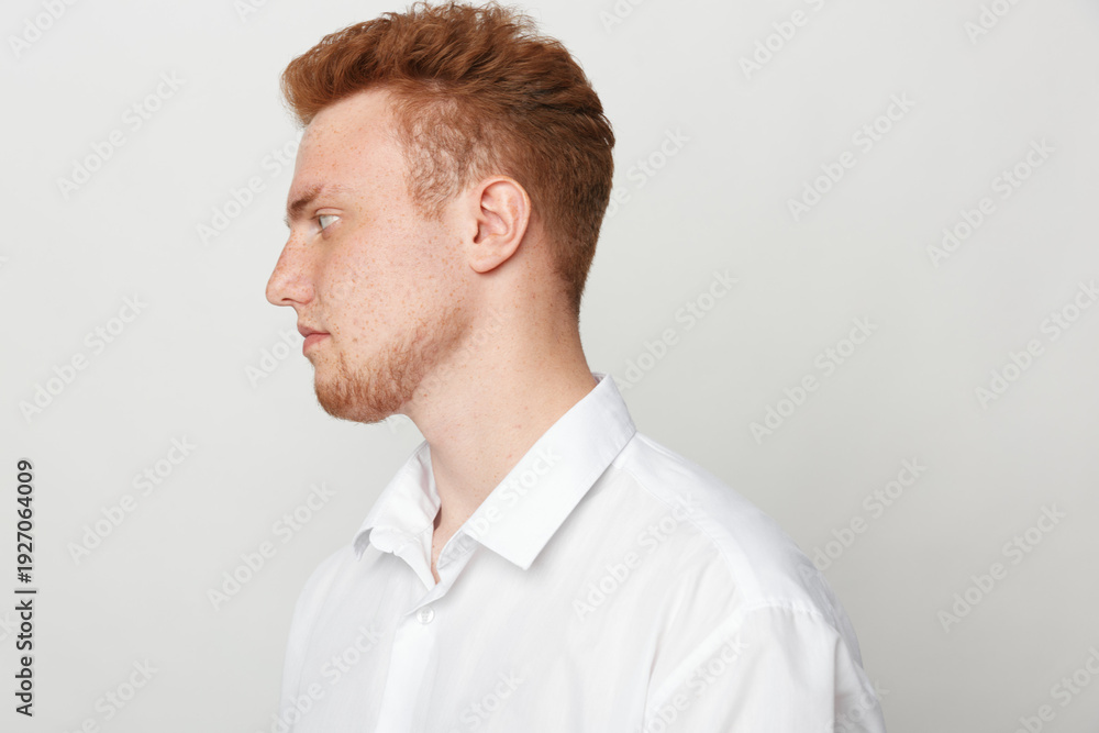 Naklejka premium Profile of a young redhead man with beard wearing white shirt looking to the left on a plain light background. Concept of casual style, portrait, and thoughtful expression.