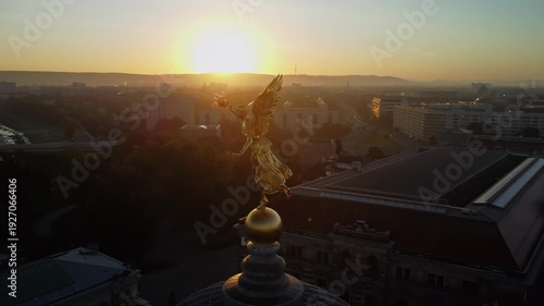 A circular aerial shoot of the statue of Pheme on the dome of the Academy of Arts in the Lipsiusbau building under sunshine at dawn with the Frauenkirche, Dresden's Old Town and the River Elbe behind 