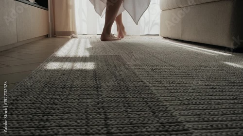 Barefoot woman walking across a rug in a modern apartment. Close-up of feet, calm home lifestyle scene in a minimalist residential interior. Neutral color palette, modern furniture in a soft morning