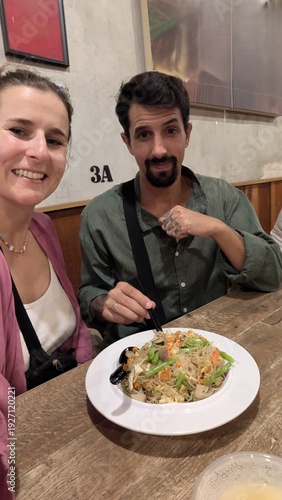 Multiracial couple with arabic man and caucasian woman taking selfie together at table, friends group laughing and enjoying chinese food lunch in indoor eatery, bonding and happiness moment