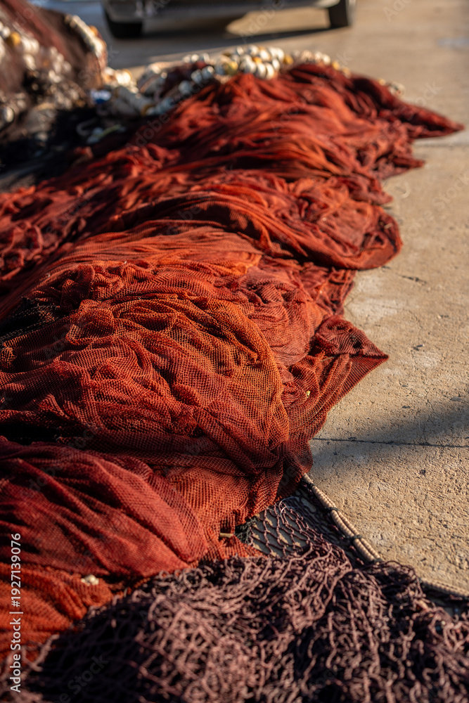 Fototapeta premium Red fishing nets drying on concrete harbor pier in warm sunlight, showing layered mesh textures, traditional maritime equipment, coastal fishing industry atmosphere, and weathered materials